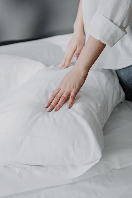 Close-up of a woman's hands adjusting a white pillow in a neatly made bed.